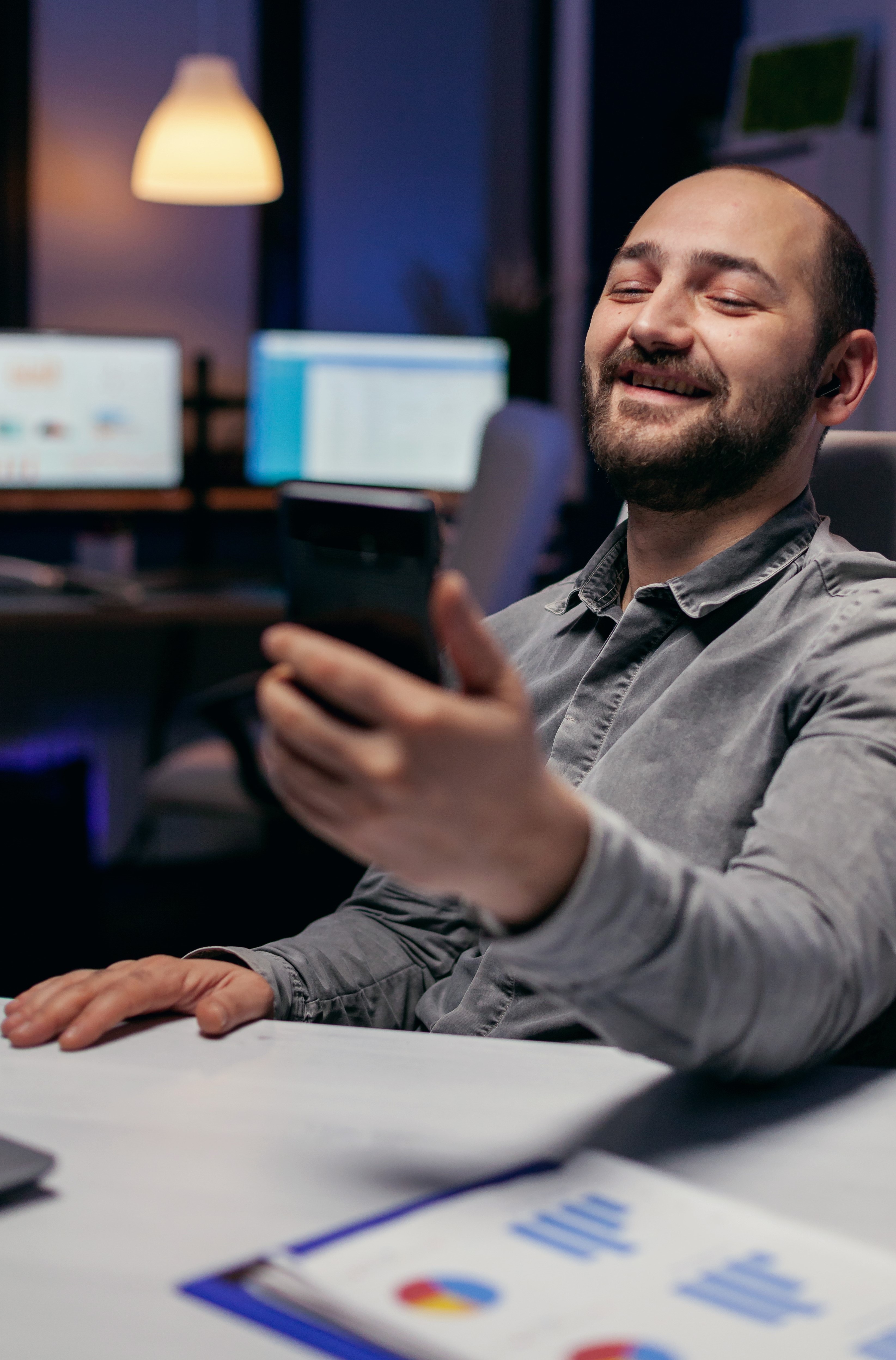 emp1 Cheerful entrepreneur saying hello talking on video call using phone. Businessman in the course of an important video conference while doing overtime at the office.